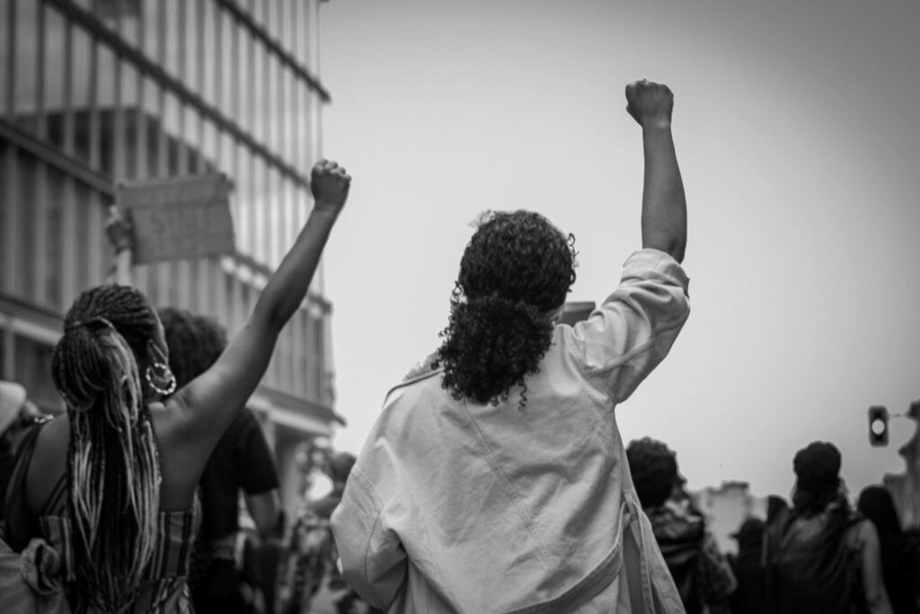 foto in bianco e nero - in una manifestazione due ragazze una accanto all'altra. Una di colore dalla capigliatura rasta, indossa una canotta e tiene il braccio verso il cielo con il pugno chiuso. L'altra è riccia con i capelli raccolti in una cosa. Ha l'impermeabile slacciato e il braccio teso verso l'alto con il pugno chiuso.