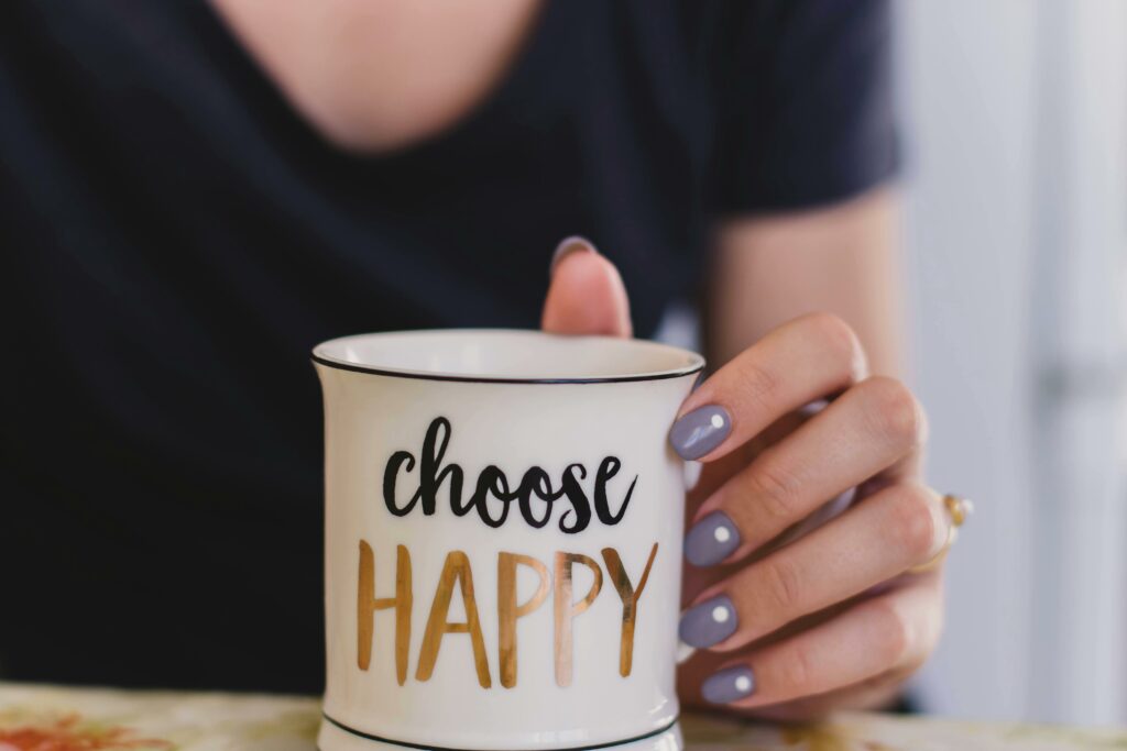 scegliere la felicità - una ragazza con la t-shirt nera dallo scollo a V ha la mano sinistra poggiata su un tavolo e con essa tiene una tazza con su scritto "choose happy". Ha lo smalto color carta da zucchero e un anello all'anulare.