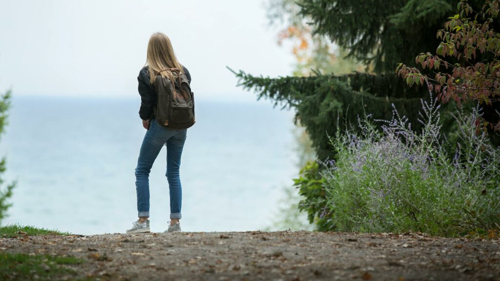 ragazza che, in un contesto campestre, guarda il mare all'orizzonte con uno zaino in spalla